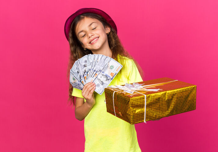 孤立的可爱的白人小女孩 戴着紫色派对帽 手里拿着礼盒 粉红色的墙上贴着钱 还有复印空间钱女孩小的