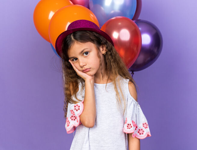 手悲伤的白人小女孩 戴着紫色的派对帽 手放在脸上 站在氦气球前面 氦气球被隔离在紫色的墙上 留着复制空间氦聚会悲伤