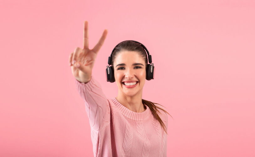 立体声戴着无线耳机听音乐的年轻女子穿着粉色毛衣微笑着摆出粉色的姿势女性科技女士