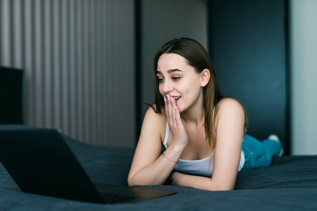 科技一个在床上使用笔记本电脑的震惊的随意的年轻女子的肖像不苟言笑电脑惊讶