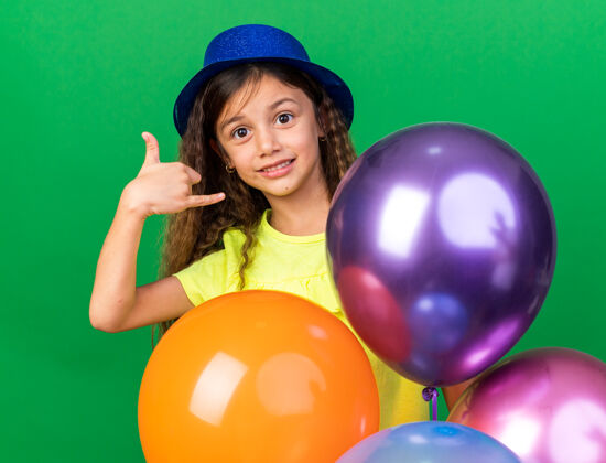 气球可爱的白人小女孩 戴着蓝色的派对帽 手里拿着氦气球 在绿色的墙壁上做着一个独立的姿势 留着复制空间请手势松散