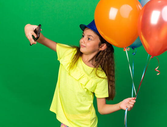 帽子可爱的白人小女孩戴着蓝色派对帽 手里拿着氦气球 在电话上自拍 被隔离在绿色的墙上 还有复印空间气球氦白种人