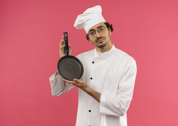 平底锅看着镜头年轻的男厨师穿着厨师制服 戴着眼镜拿着煎锅厨师制服烹饪