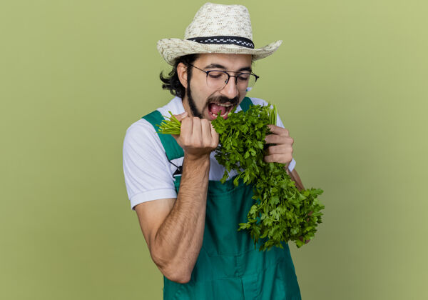衣服年轻的留着胡须的园丁 穿着连体衣 戴着帽子 手里拿着新鲜的草药 站在淡绿色的墙上吃着它人新鲜人