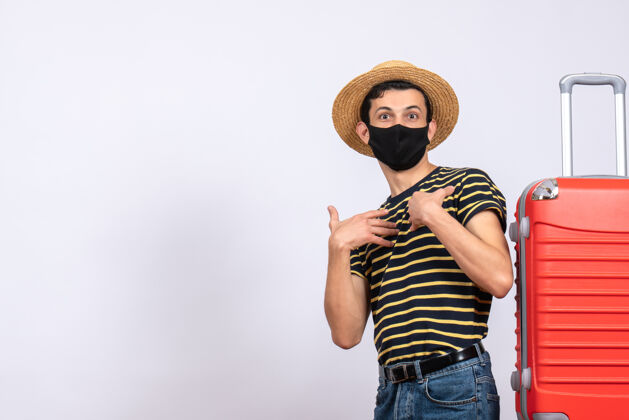 男正面图：戴着黑色面具的年轻游客站在红色手提箱旁指着自己男站年轻的游客