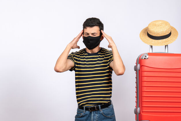 视图正面图：体贴的年轻游客 戴着黑色面具 双手抱着头站在红色手提箱旁人面具红色