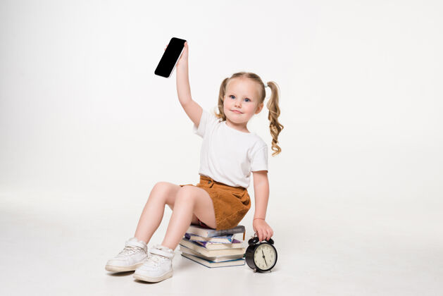 可爱一个小女孩拿着手机屏幕坐在一堆书上的画像教育美丽女学生