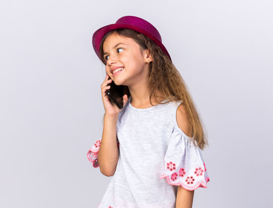 壁板微笑着戴着紫色派对帽的白人小女孩一边看一边讲着电话 隔离在白色的墙上 留着复印空间生日孤立聚会