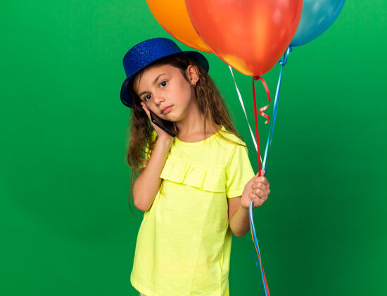 气球失望的白人小女孩 戴着蓝色派对帽 手里拿着氦气球 在绿色墙壁上讲着电话 还有复印空间女孩举行孤立