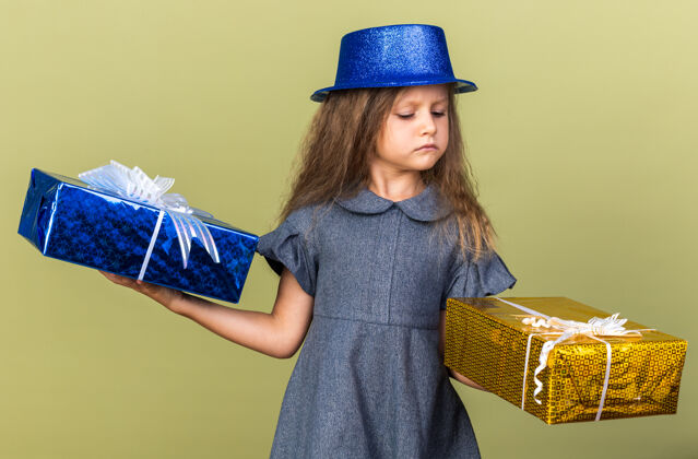 小恼怒的金发小女孩拿着蓝色的派对帽 看着橄榄绿墙上的礼品盒 还有复印空间金发孤立帽子