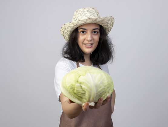 女性微笑着的年轻黑发女园丁穿着制服 戴着园艺帽 把卷心菜隔离在白墙上人年轻人脸