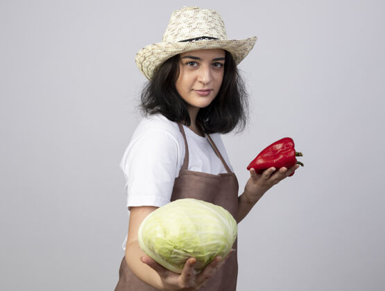 表情自信的年轻黑发女园丁穿着制服 戴着园艺帽 把卷心菜和红辣椒隔离在白墙上人卷心菜人