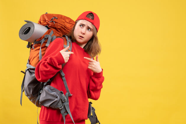 手持正面图穿着红色背包的年轻女旅行者指着自己帽子指点时尚
