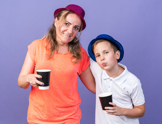 男孩高兴的小斯拉夫男孩拿着蓝色的聚会帽拿着纸杯站着 母亲戴着紫色的聚会帽孤立地站在紫色的墙上 留着复印空间斯拉夫人帽子杯子