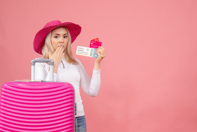 拿着正面图：站在粉红色手提箱后面拿着卡片和票的年轻女士成人站着票