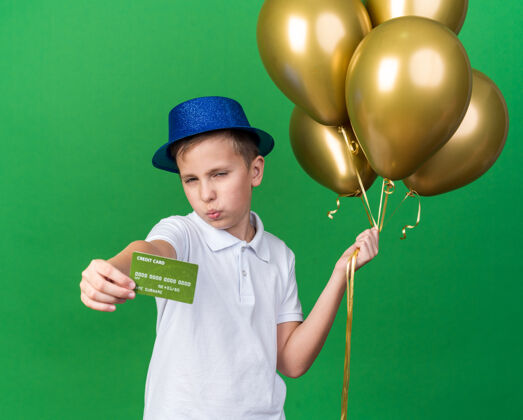 氦自信的年轻斯拉夫男孩 戴着蓝色派对帽 手里拿着氦气球 信用卡被隔离在绿墙上 留着复印空间信用持有孤立
