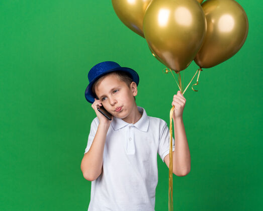 帽子自信的年轻斯拉夫男孩 戴着蓝色派对帽 手里拿着氦气球 一边打电话 一边看着隔离在绿色墙壁上的一面 一边留着复印空间年轻男孩电话