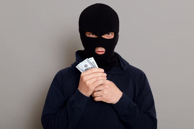 面具愤怒的窃贼用伪造的表情看着前面 手里拿着赃款严重灰色背景盗窃