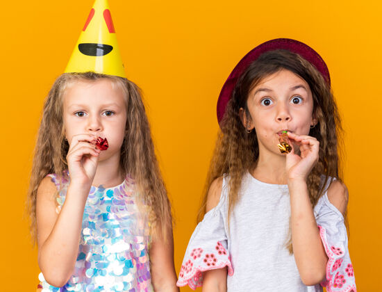 小快乐的小漂亮女孩 戴着派对帽吹着口哨 隔离在橘黄色的墙上 还有复制空间漂亮吹生日