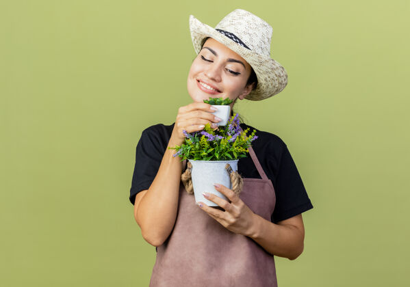 快乐年轻漂亮的女园丁 围着围裙 戴着帽子 手里拿着盆栽植物 站在淡绿色的墙上 面带微笑地看着它微笑姿势人