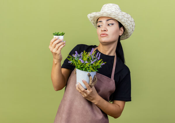 感觉年轻漂亮的女园丁 围着围裙 戴着帽子 手里拿着盆栽植物 站在淡绿色的墙上 看起来很困惑 试图做出选择选择姿势人