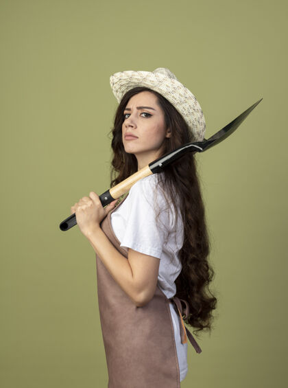 橄榄自信的年轻女园丁身着制服 戴着园艺帽 站在一旁 手持铁锹 孤零零地站在橄榄绿的墙上女性信心黑桃