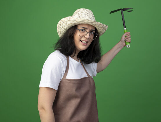 女人恼怒的年轻黑发女园丁戴着眼镜和制服戴着园艺帽拿着锄头耙隔离在绿色的墙上眼镜姿势制服