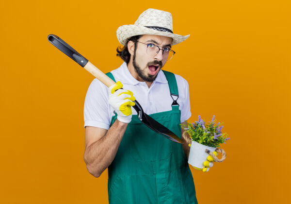人年轻的留着胡子的园丁 穿着连体衣 戴着帽子 拿着铲子和盆栽植物 站在橙色的墙上 看上去很困惑衣服脸年轻人