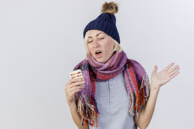人一个戴着冬日帽 戴着围巾的年轻金发病女人站在那里 举起手 手里拿着一包隔离在白墙上的药片金发包装药丸
