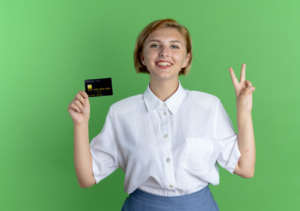 绿色年轻的微笑金发俄罗斯女孩持有信用卡和手势胜利手势手微笑背景