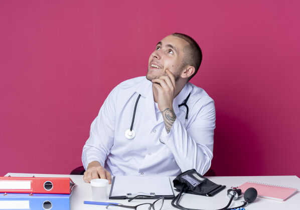 男性面带微笑的年轻男医生穿着医用长袍 听诊器坐在办公桌旁 手放在下巴上 孤零零地看着粉红色的墙上人公民粉色