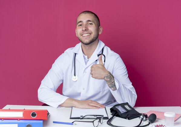 工作面带微笑的年轻男医生穿着医用长袍 听诊器坐在办公桌旁 工作工具上的拇指被隔离在粉红色的墙上拇指人桌子