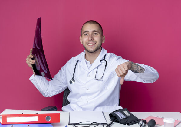指点面带微笑的年轻男医生 穿着医用长袍 手持听诊器坐在办公桌旁 手里拿着工作工具 拿着x光片 指着隔离在粉红色墙上的墙按住X光姿势