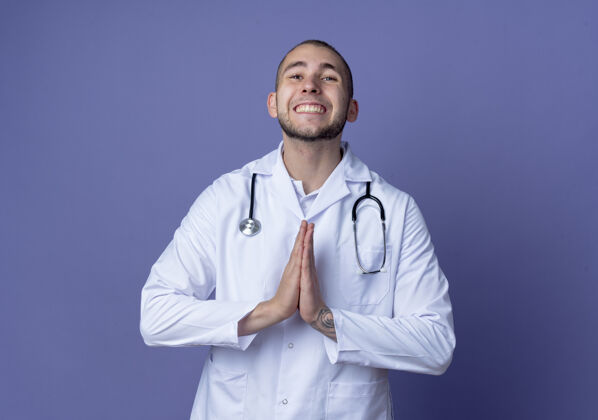 手面带微笑的年轻男医生 身穿医用长袍 脖子上戴着听诊器 双手放在紫色的墙上做着祈祷的姿势穿着姿势医学
