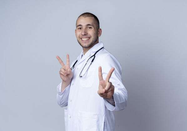 医学面带微笑的年轻男医生 身穿医用长袍 脖子上戴着听诊器 站在侧视图中 在白墙上做着隔离的和平标志人人表情