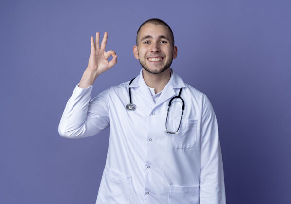 听诊器微笑着的年轻男医生穿着医用长袍 脖子上戴着听诊器 在紫色的墙上做着“ok”的手势年轻紫色医学