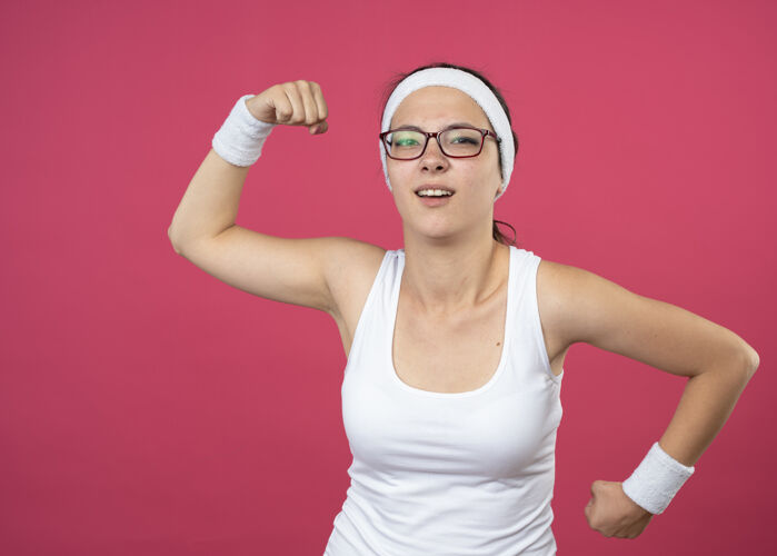 运动戴着眼镜 戴着头带和腕带的恼怒的运动型年轻女子上下握拳 孤立地站在粉红色的墙上女人头带恼怒