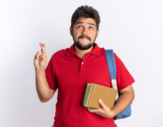 抱着年轻的留着胡子的学生 穿着红色马球衫 背着背包 拿着笔记本 在白色的背景上 用希望的表情交叉着手指许愿做希望背包