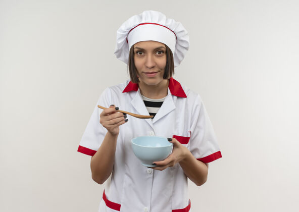 脸高兴的年轻女厨师在厨师制服举行汤匙和碗隔离在白墙上人衣服人