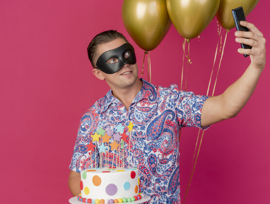 拿着高兴的年轻人戴着化装眼罩 拿着气球和蛋糕 在粉红色上自拍年轻眼睛自拍