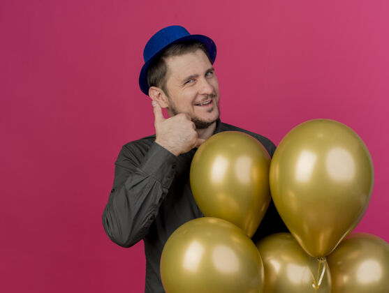 蓝色高兴的年轻人戴着蓝色的帽子站在气球旁边 显示电话的手势孤立的粉红色男人粉色站着