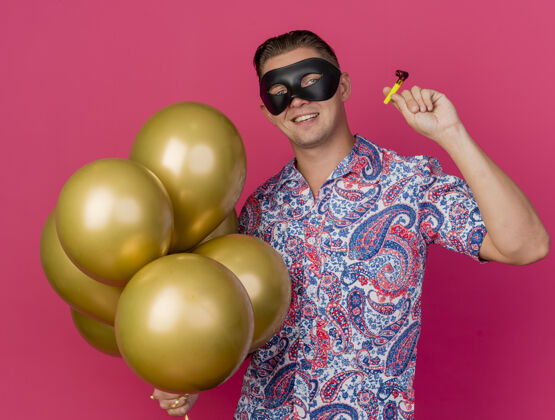 穿面带微笑的年轻人戴着化装眼罩 手里拿着气球 吹着粉红色的派对风吹风机化装舞会面具