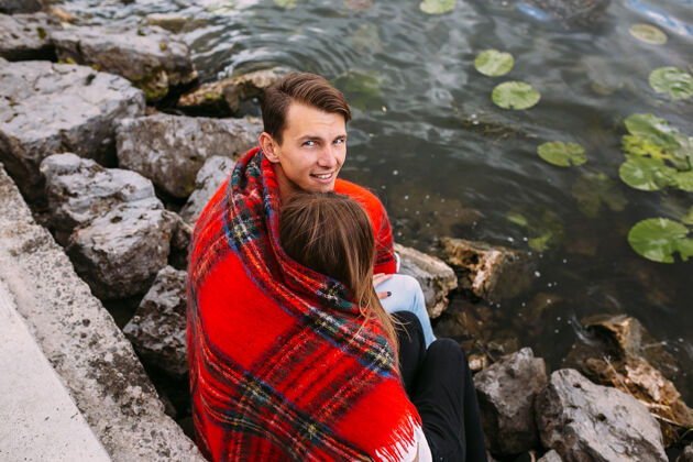年轻美丽的年轻夫妇坐在石堤上 裹着毯子男人拥抱情侣