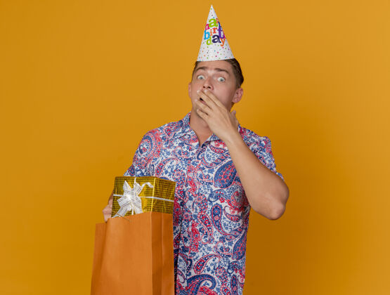 小伙子害怕的年轻人戴着生日帽拿着礼物包 用手遮住脸上的橙色隔离橘子戴着聚会