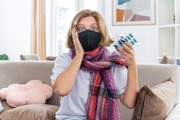 房间不健康的年轻女子 脖子上围着暖和的围巾 戴着面膜 感觉不舒服 生病 患流感 吃感冒药 坐在客厅的沙发上 看起来很困惑流感围巾感冒