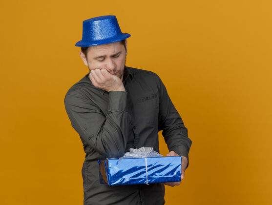 戴着悲伤的年轻人戴着蓝色的帽子拿着礼物盒把肘部孤立在橙色蓝色礼物派对