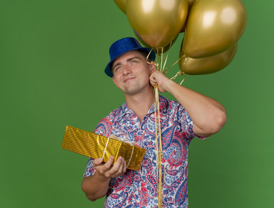 盒子高兴的年轻人戴着蓝色帽子拿着气球与礼品盒隔离在绿色礼物拿着派对