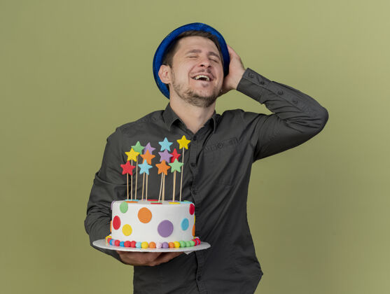 戴着微笑着闭上眼睛的年轻人穿着黑色衬衫 戴着蓝色帽子拿着蛋糕 手放在橄榄绿上孤立的帽子上眼睛橄榄色黑色
