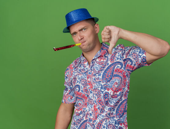 不愉快一个戴着蓝帽子的年轻人在派对上吹着吹风机 大拇指朝下 孤零零地站在草地上派对帽子吹