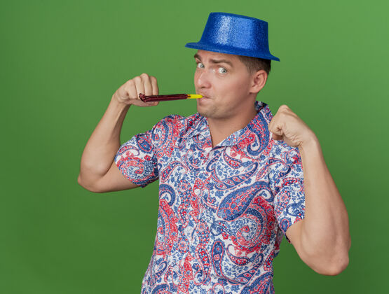 男高兴的年轻人戴着蓝色的帽子吹聚会吹风机显示出强大的姿态孤立的绿色吹年轻绿色